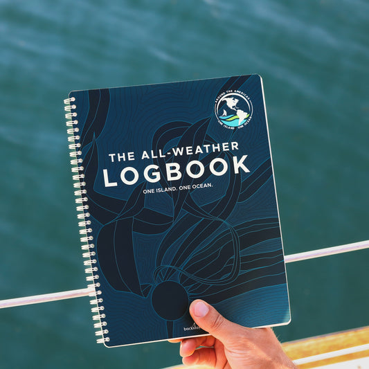 All-Weather Logbook with One Island One Ocean cover design featuring kelp and expedition logo on a spiral-bound book resting on calm water.