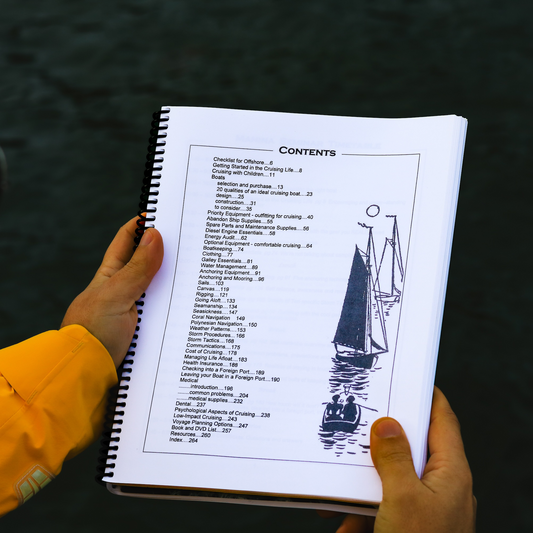 Spiral bound Offshore Cruising Companion book open to the table of contents page, held over water in foul weather sailing gear.