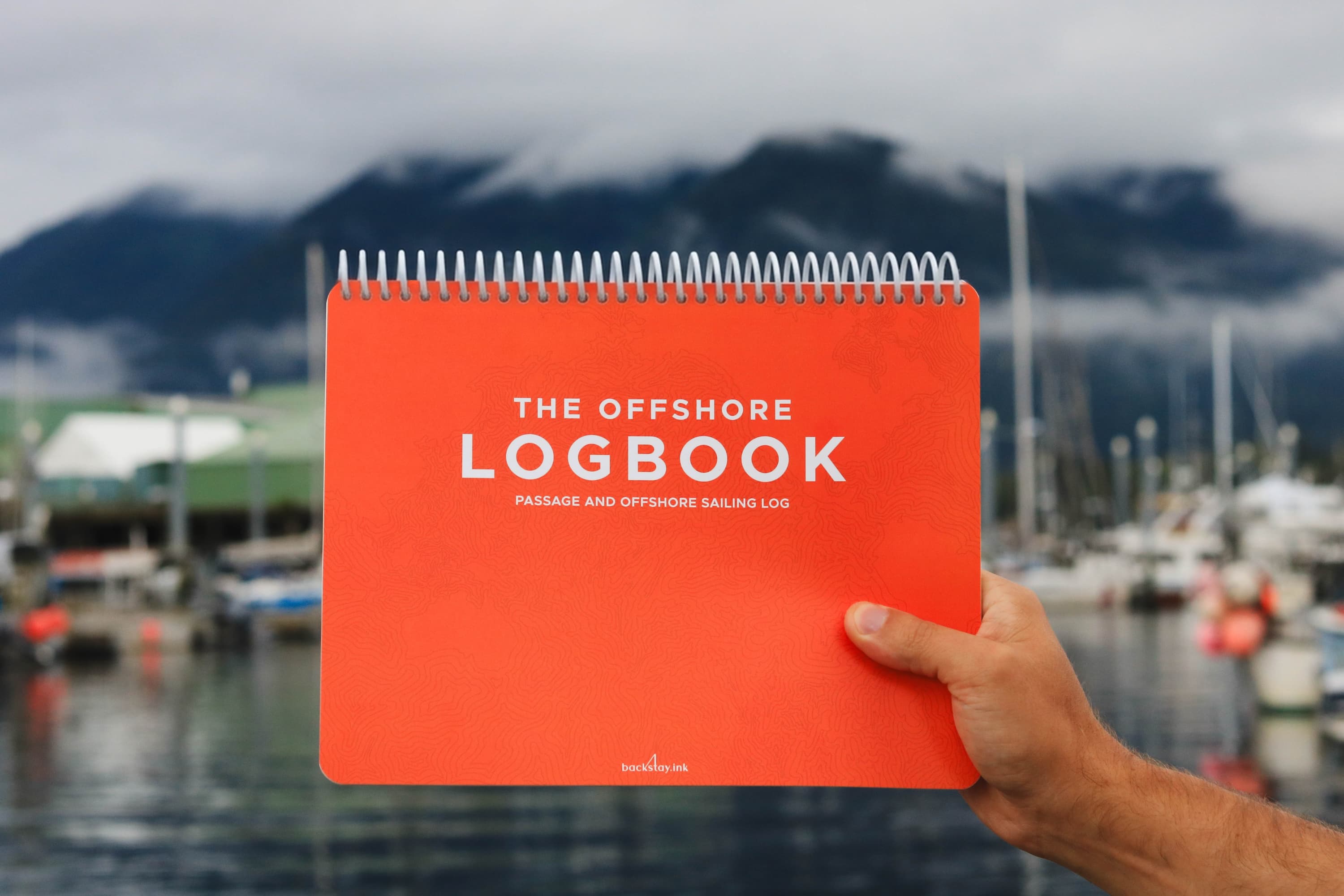 The Offshore Logbook held by a sailor with a marina and mountains in the background.

