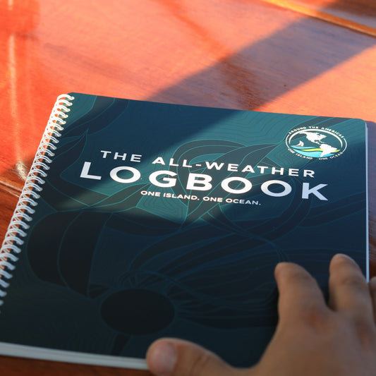 All-Weather Logbook with One Island One Ocean cover design featuring kelp and expedition logo on a spiral-bound book resting on calm water.