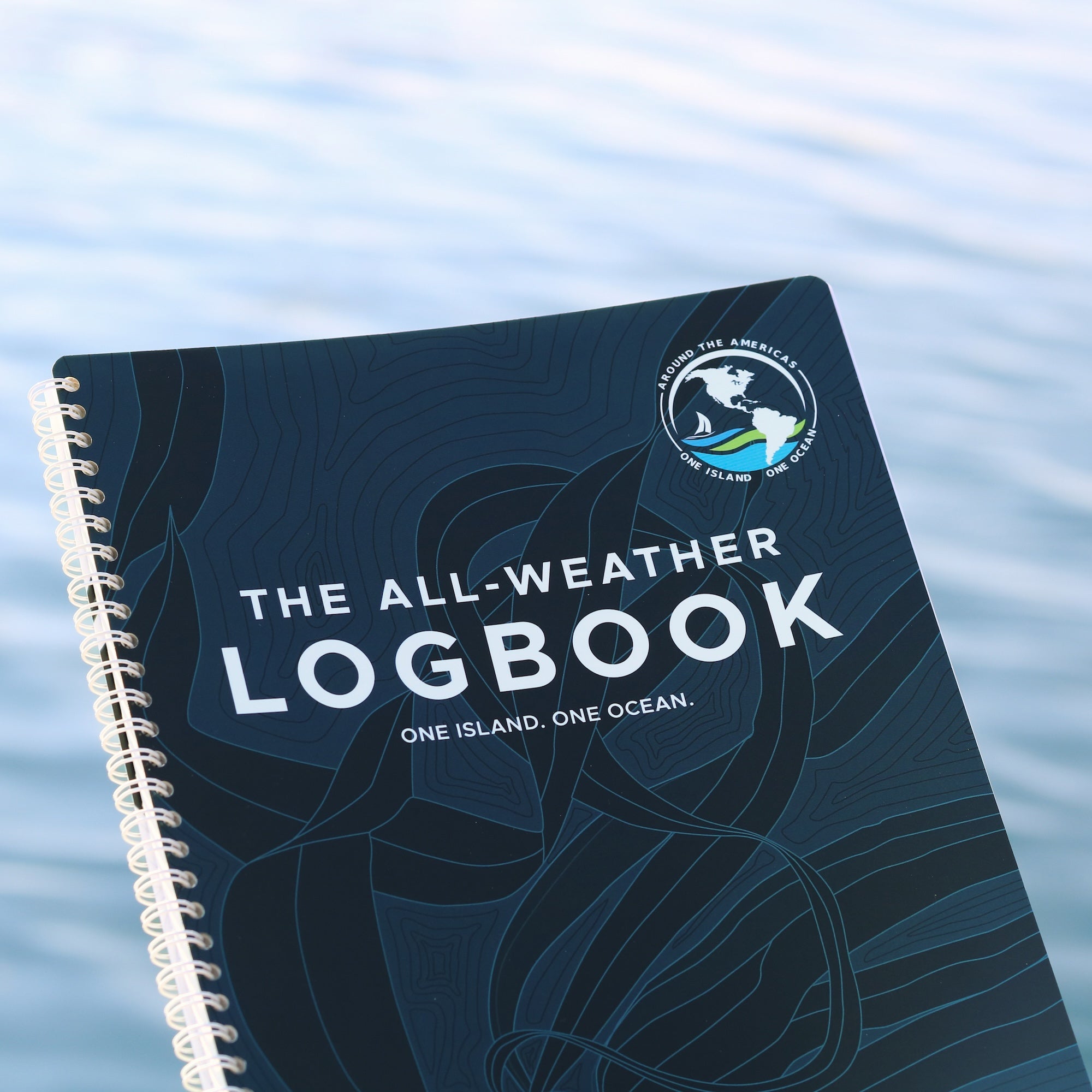 All-Weather Logbook with One Island One Ocean cover design featuring kelp and expedition logo on a spiral-bound book resting on calm water.