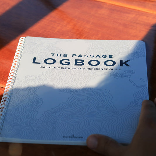 Hand holding the Backstay Passage Logbook on a wooden surface with spiral binding visible.