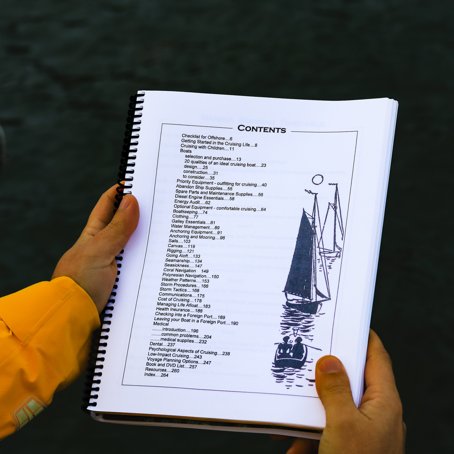 Spiral bound Offshore Cruising Companion book open to the table of contents page, held over water in foul weather sailing gear.
