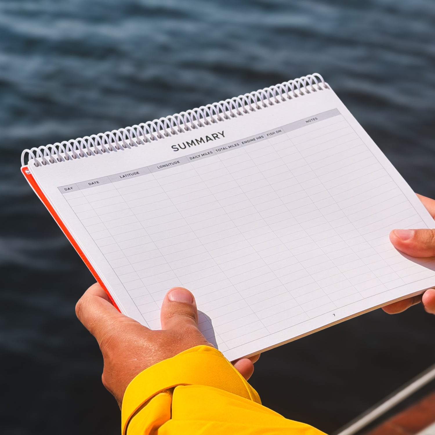 A sailor holding a spiral-bound summary page in the Offshore Logbook, photographed outdoors with water in the background.